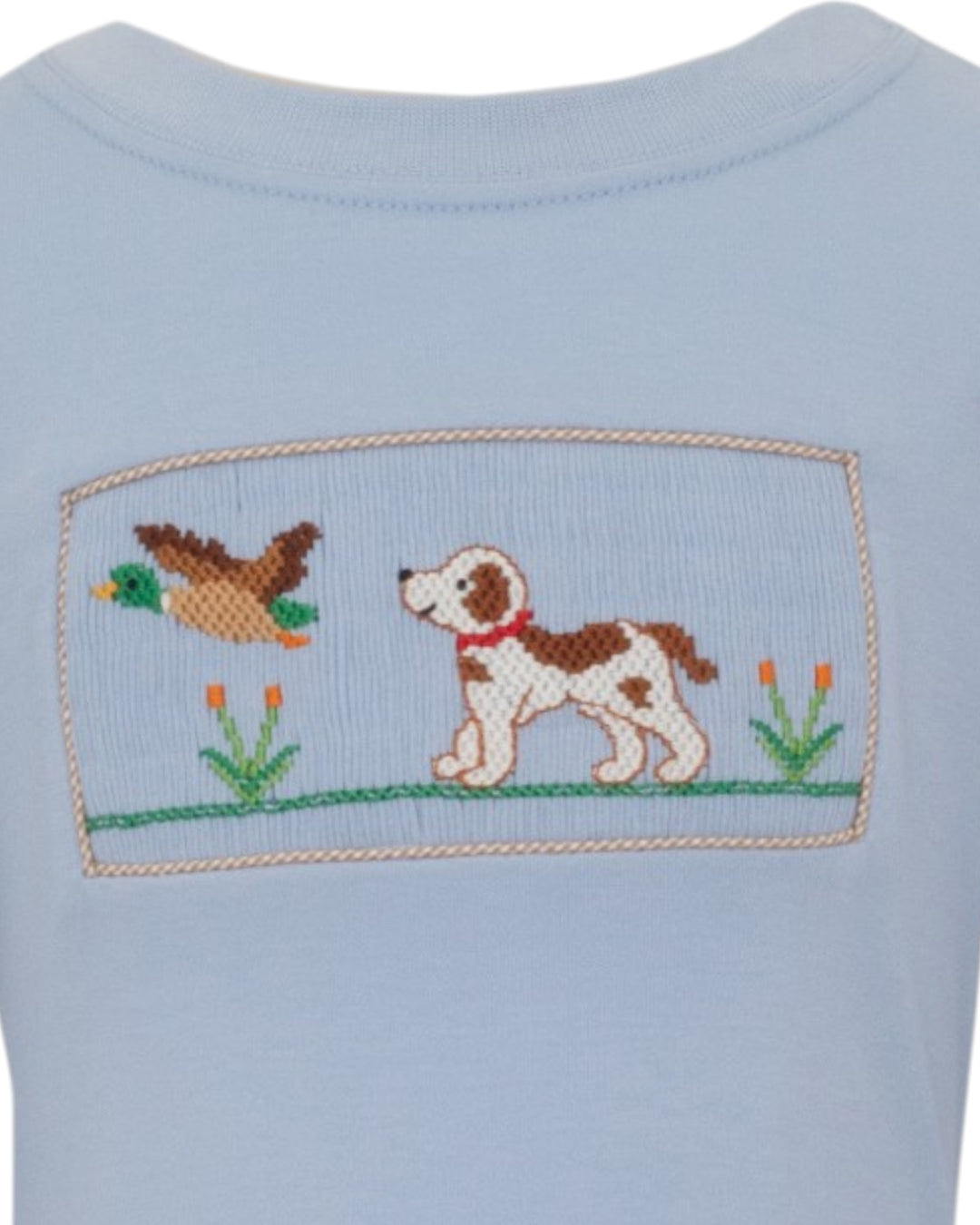 Close-up of a light blue garment featuring a cross-stitch style patch with a brown and white dog, a duck, and small flowers, that store