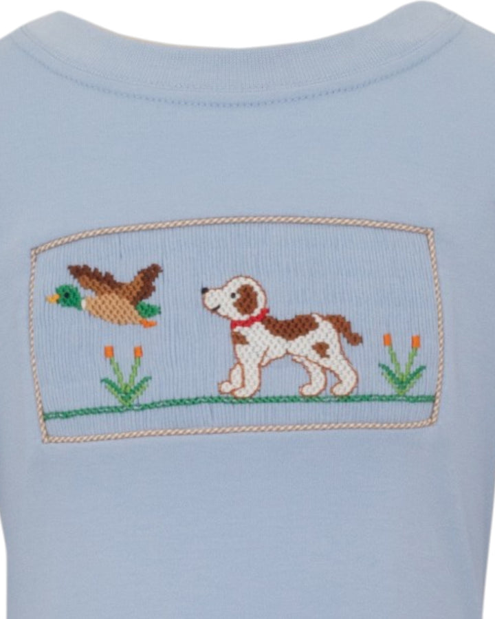 Close-up of a light blue garment featuring a cross-stitch style patch with a brown and white dog, a duck, and small flowers, that store