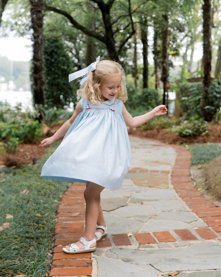 Young girl in a light blue dress with a white bow standing on a stone path in a garden.