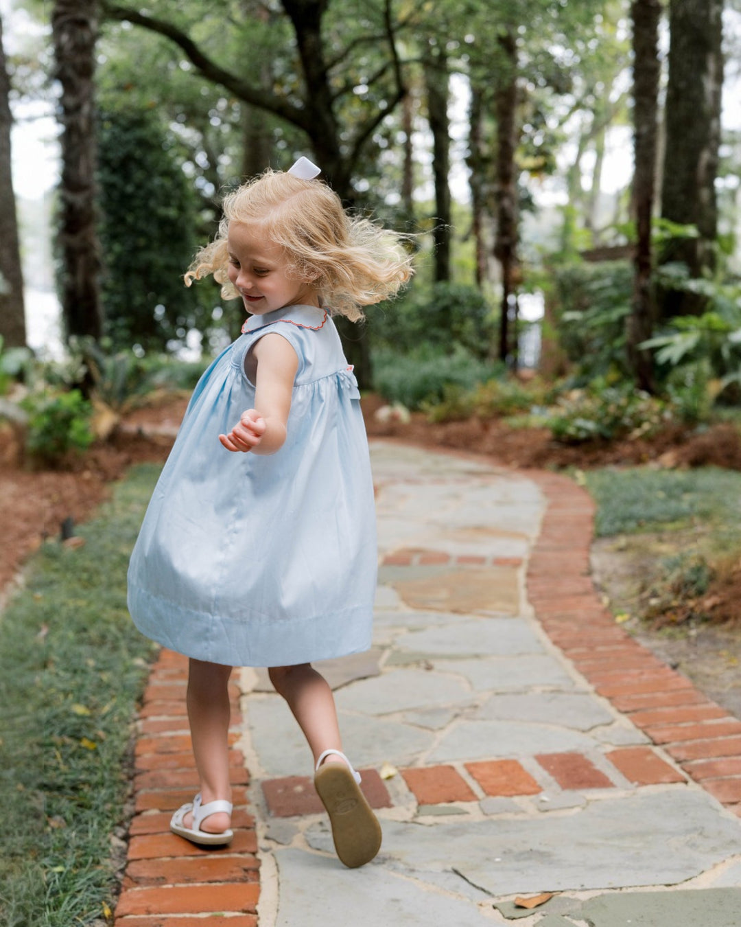 Child in a light blue dress walking on a stone path in a garden.