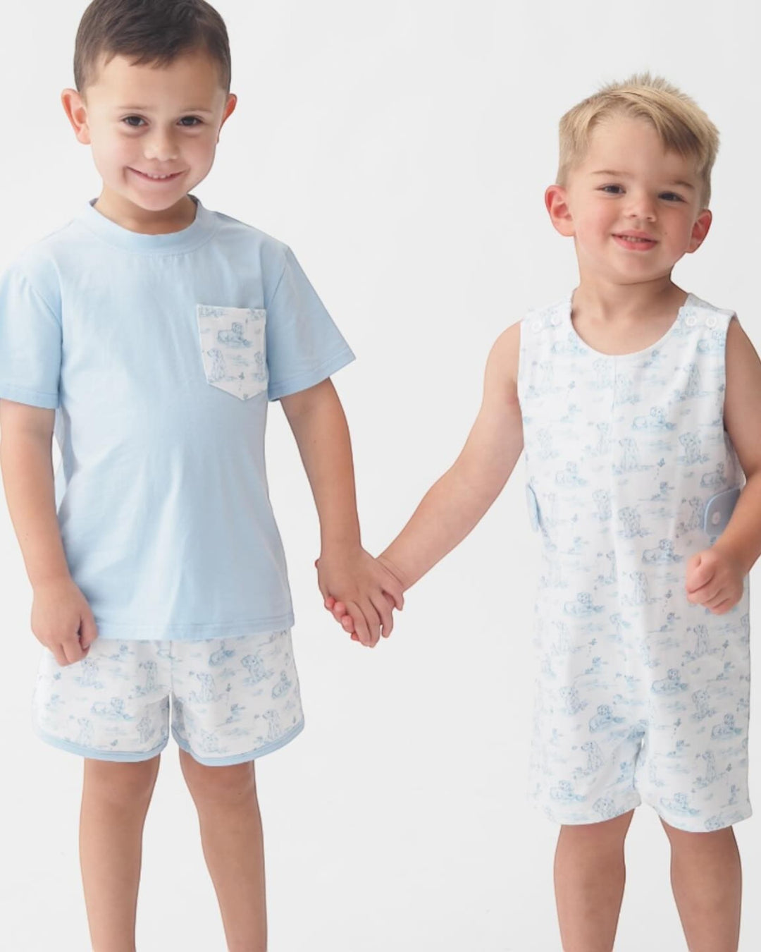 Two young boys holding hands, wearing matching light blue outfits with subtle patterns on a white background.