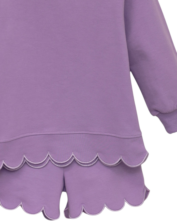A close-up of a lavender purple long-sleeve top or dress with a scalloped hem, paired with matching scalloped shorts, shown against a plain white background, that store