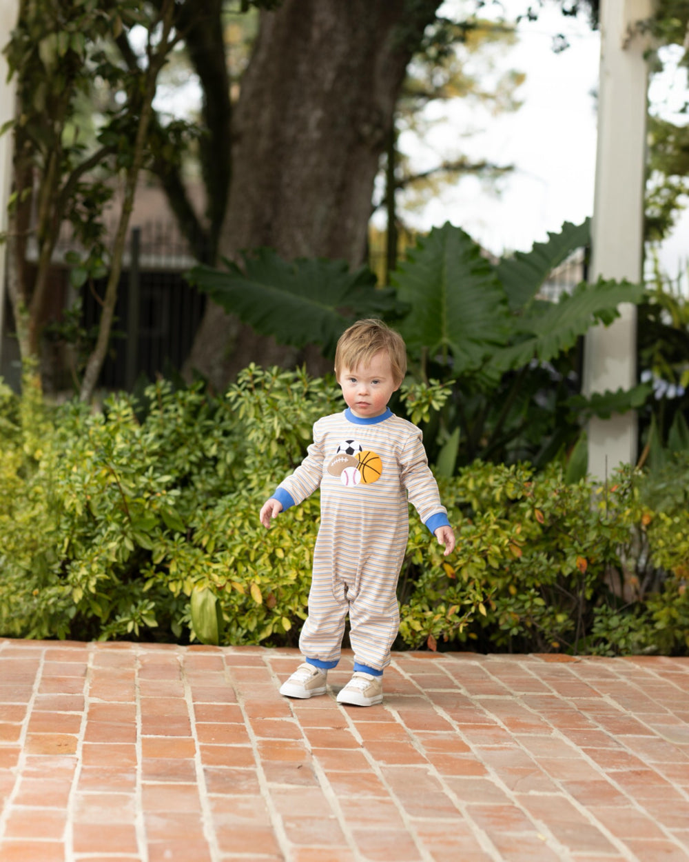 Child in a patterned onesie standing on a brick path with greenery in the background