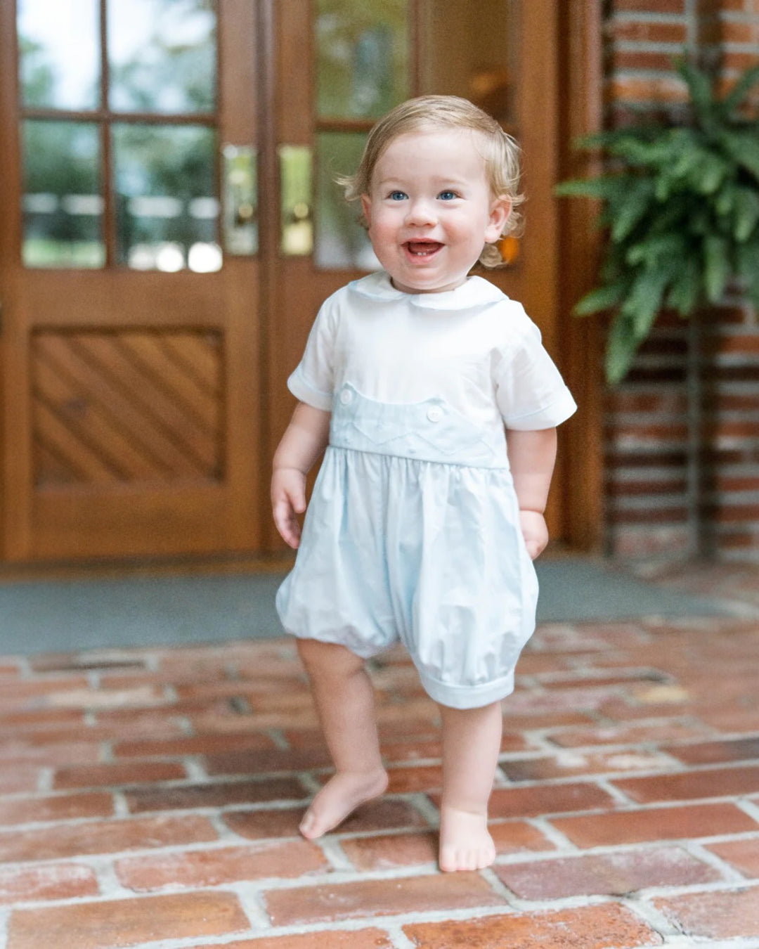 Child in a light blue dress standing on a brick floor with a wooden door and plant in the background.
