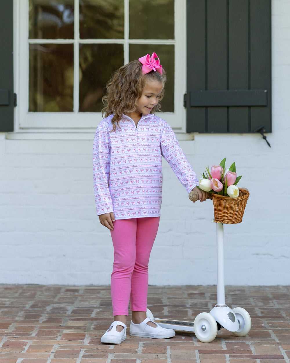A young girl stands on a white scooter in front of a white brick wall and dark shutters, wearing a light purple and white patterned long-sleeve quarter-zip top, bright pink leggings, and white shoes. She has a pink bow in her hair and is holding a small basket of tulips, that store