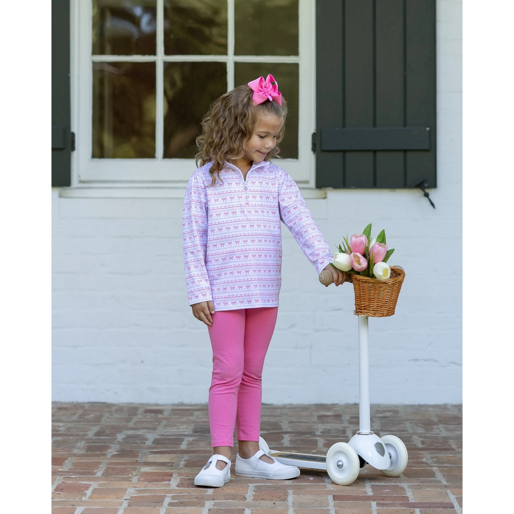 A young girl stands on a white scooter in front of a white brick wall and dark shutters, wearing a light purple and white patterned long-sleeve quarter-zip top, bright pink leggings, and white shoes. She has a pink bow in her hair and is holding a small basket of tulips, that store