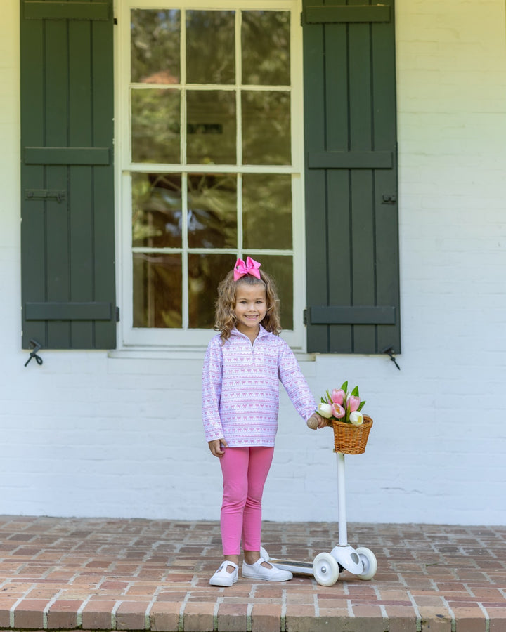 A young girl stands on a brick porch in front of a white house with dark green shutters, wearing a light purple patterned long-sleeve top, bright pink leggings, and white sneakers. She is holding a small basket on a white scooter with pink tulips, that store