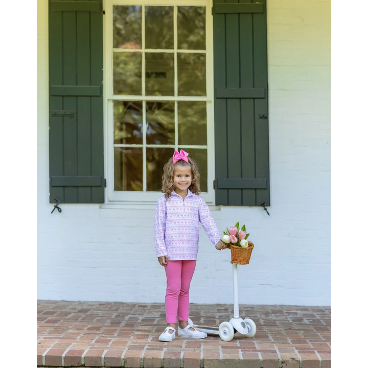 A young girl stands on a brick porch in front of a white house with dark green shutters, wearing a light purple patterned long-sleeve top, bright pink leggings, and white sneakers. She is holding a small basket on a white scooter with pink tulips, that store