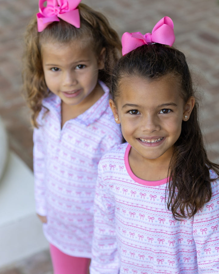 Two young girls wearing matching pink and white patterned long-sleeve shirts and large pink hair bows, photographed outdoors with a blurred neutral background, that store