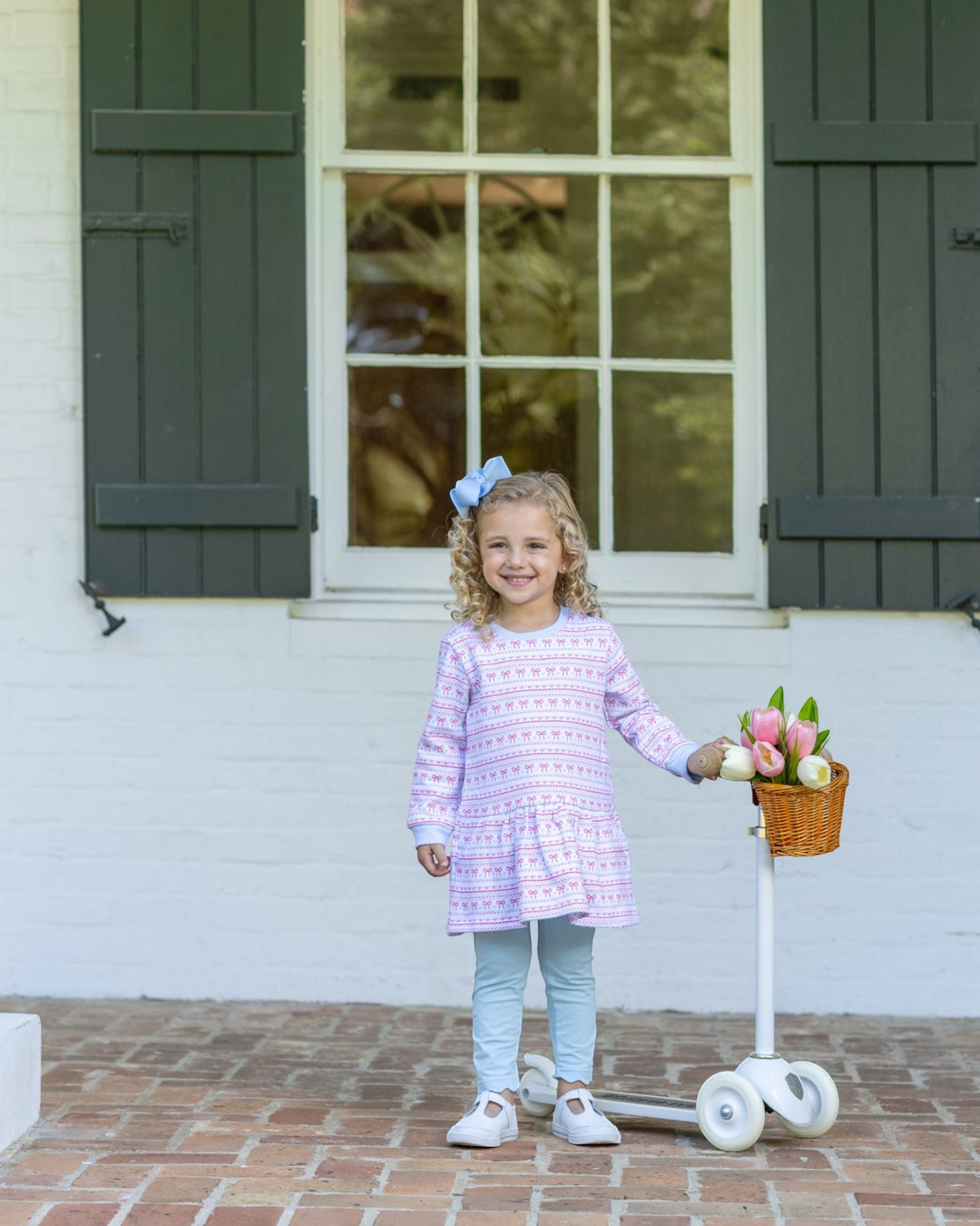 A young girl with curly blonde hair stands on a brick patio in front of a white house with dark green shutters. She is wearing a lavender and white patterned long-sleeve tunic top, light blue leggings, and white sneakers, and is holding a small wicker basket filled with pink and white flowers next to a white scooter, that store