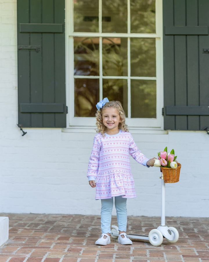 A young girl with curly blonde hair stands on a brick patio in front of a white house with dark green shutters. She is wearing a lavender and white patterned long-sleeve tunic top, light blue leggings, and white sneakers, and is holding a small wicker basket filled with pink and white flowers next to a white scooter, that store