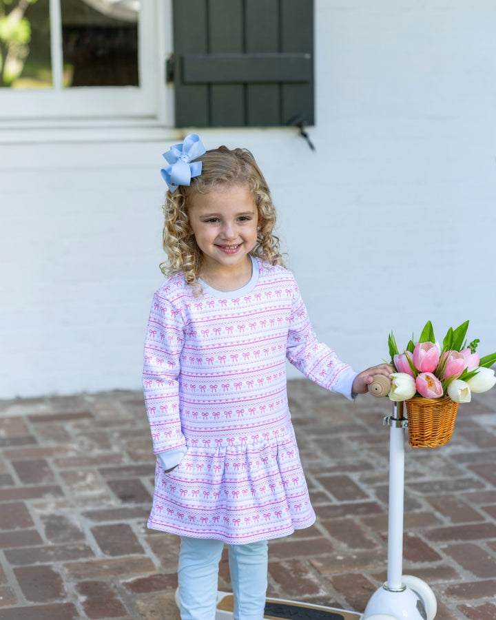 A young girl with curly blonde hair wearing a light purple and pink patterned long-sleeve dress and light blue pants, standing next to a white scooter with a basket of pink and white tulips in front of a white house, that store
