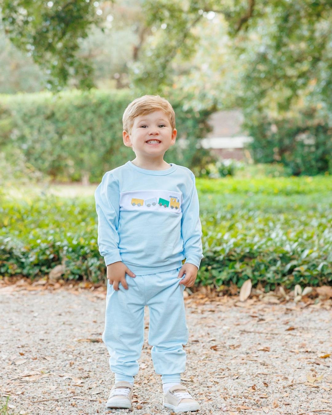 A young boy wearing a light blue two-piece pajama set stands outdoors on a gravel path with green hedges and trees in the background. The long-sleeve top has a small colorful graphic across the chest, that store