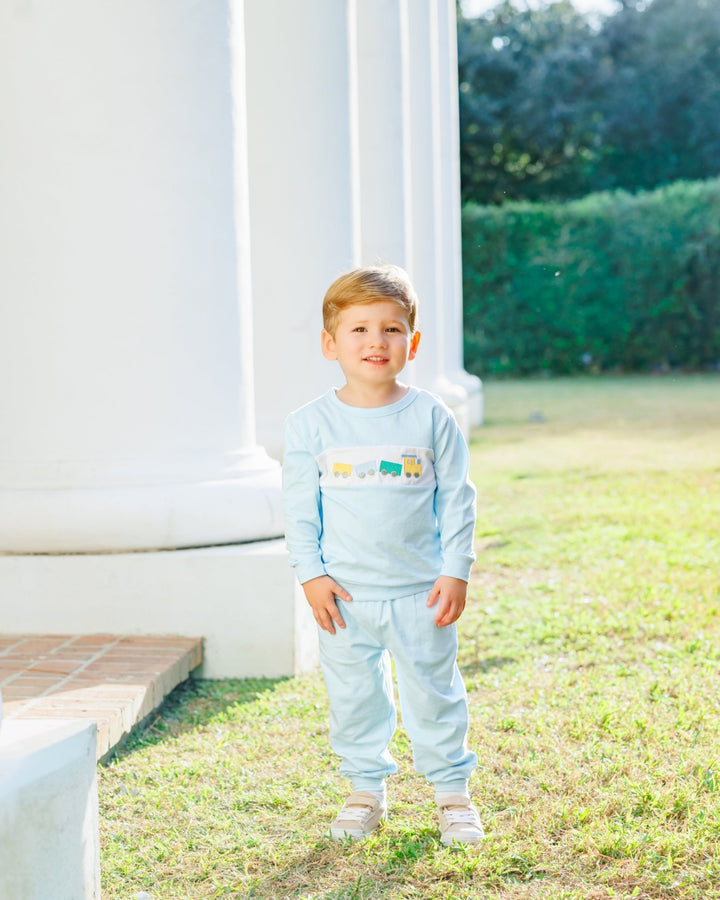 A young toddler boy wearing a light blue long-sleeve pajama set with a colorful train graphic across the chest, standing on a grassy lawn in front of a white classical column, that store