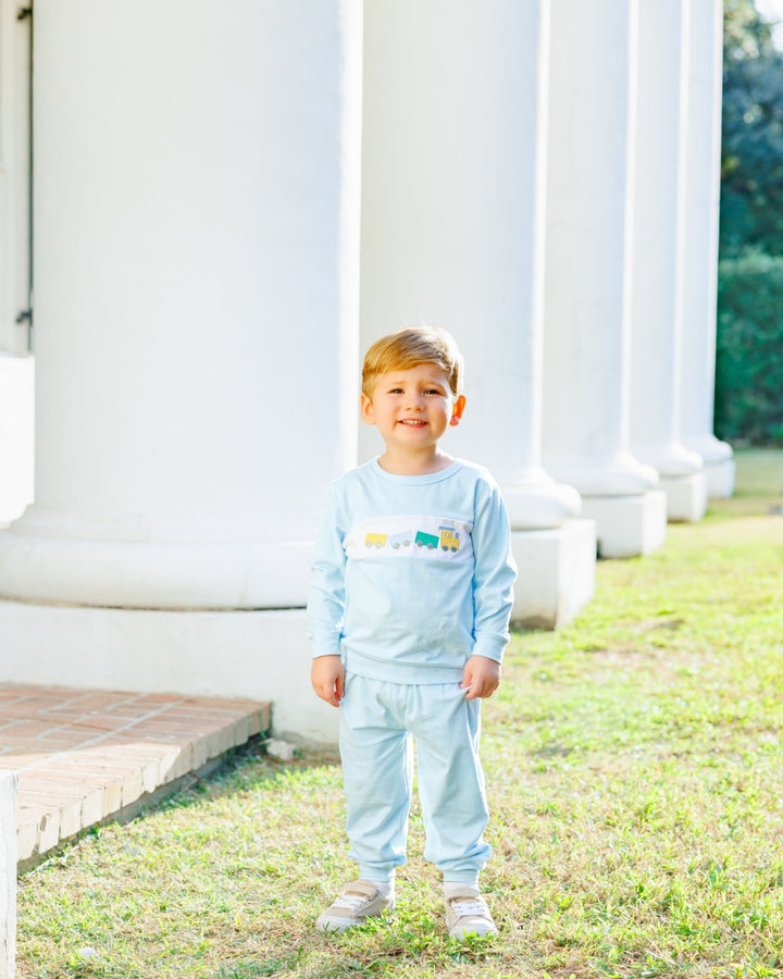A young boy stands outdoors on grass in front of large white columns, wearing a light blue long-sleeve pajama set with a colorful graphic across the chest, that store
