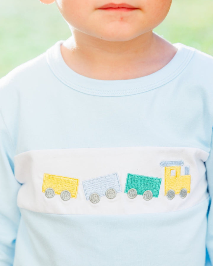 Close-up of a child wearing a light blue long-sleeve shirt with a white chest panel featuring embroidered toy trains in yellow, blue, and green, that store