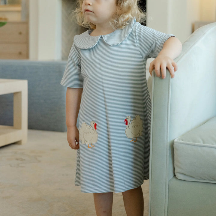 Child wearing a blue and white striped dress with duck appliqués in a living room, that store