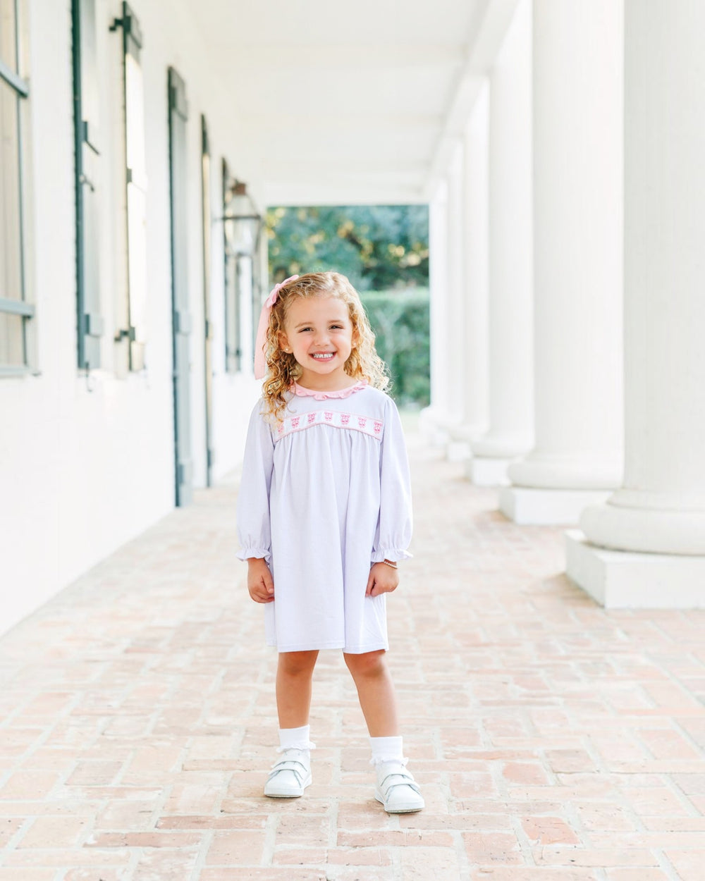 A young girl with blonde hair stands smiling on a brick walkway in front of a white building with columns, wearing a light lavender long-sleeve dress with pink embroidery across the chest, white socks, and white shoes, that store
