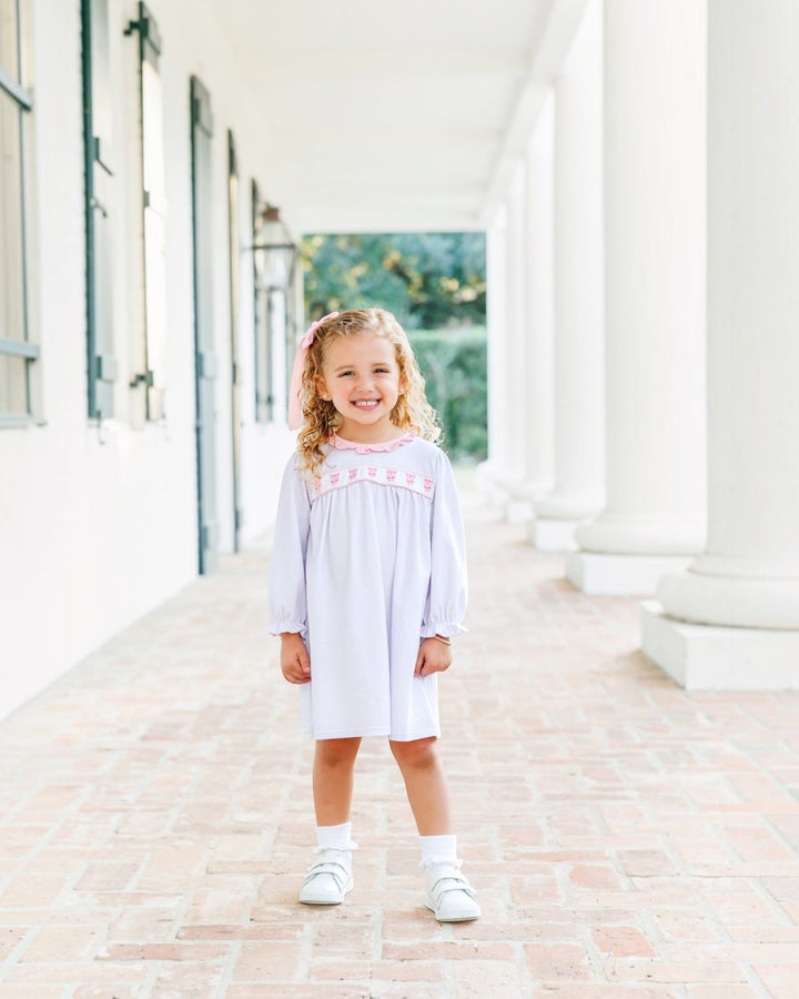 A young girl with blonde hair stands smiling on a brick walkway in front of a white building with columns, wearing a light lavender long-sleeve dress with pink embroidery across the chest, white socks, and white shoes, that store