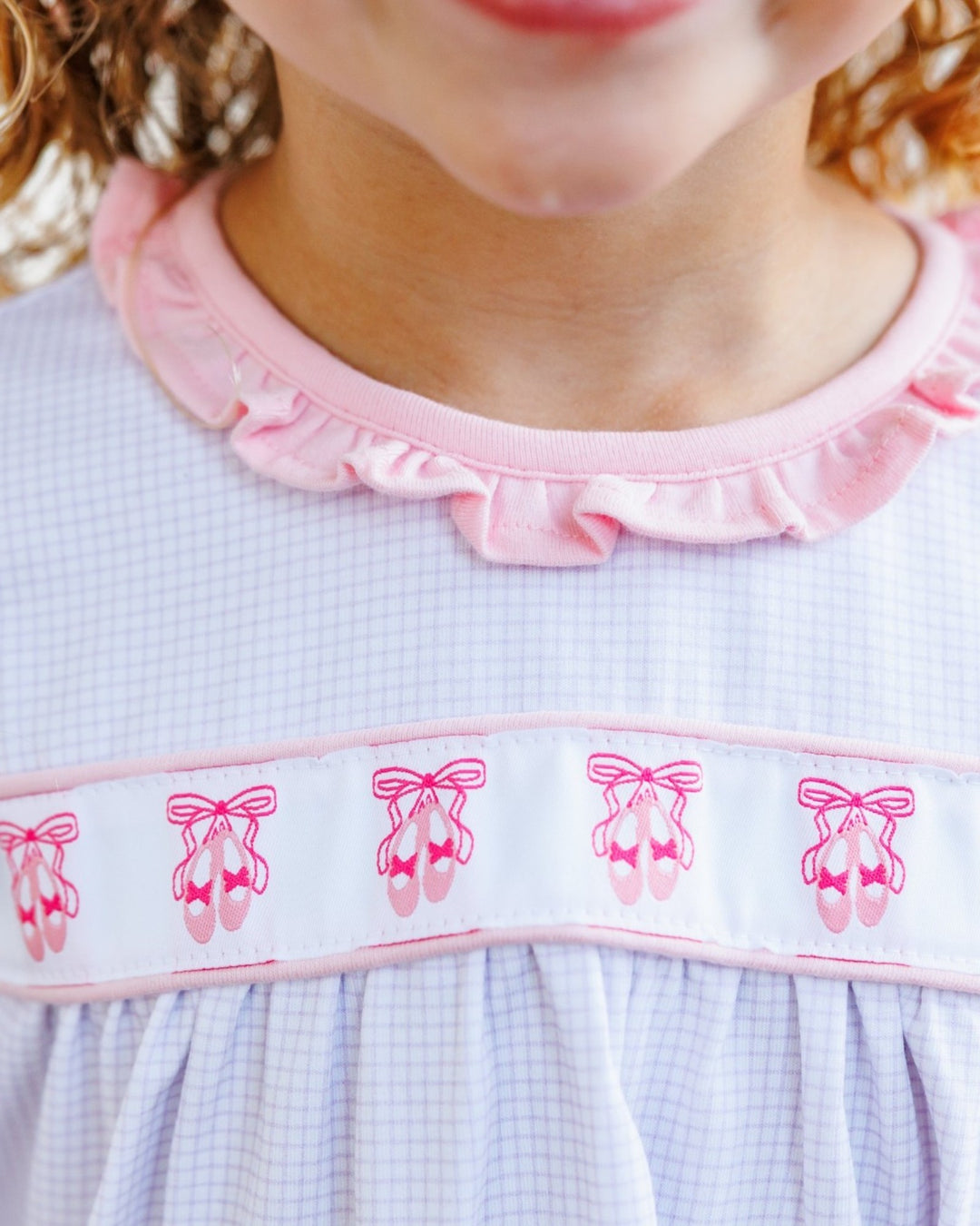 Close-up of a child wearing a light blue and white gingham dress with a pink ruffled collar and a white band decorated with pink ballet shoe motifs, that store