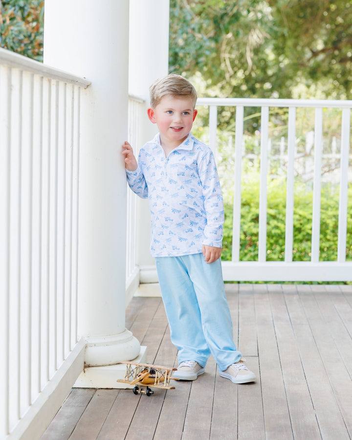 A young boy stands on a white porch wearing a light blue and white patterned long-sleeve polo shirt and matching light blue pants. A small toy airplane sits on the wooden deck floor nearby, with white railings and green foliage in the background, that store