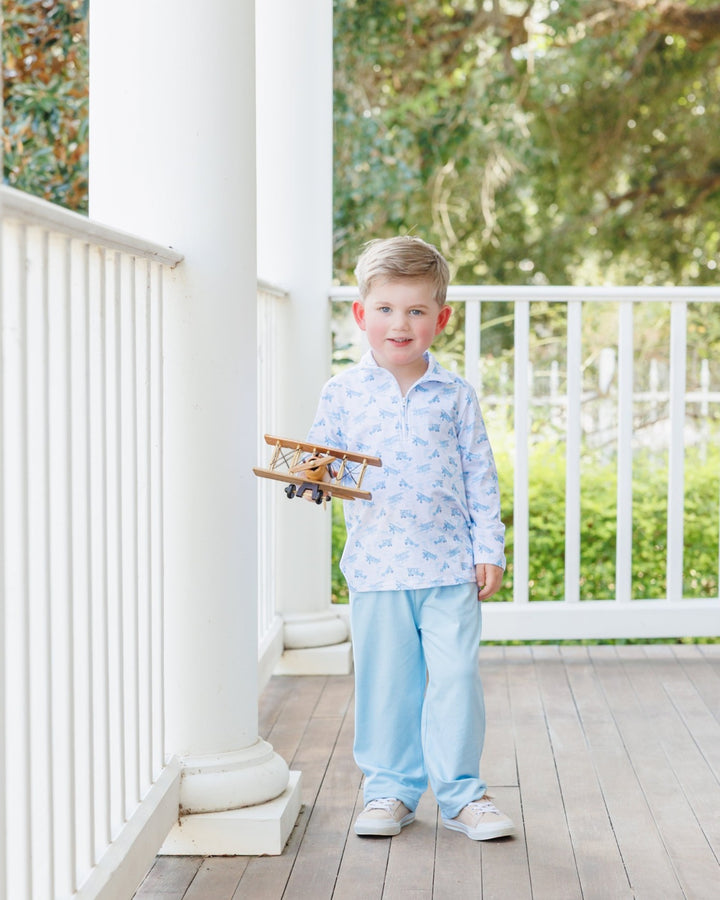 A young boy stands on a white porch wearing a light blue and white patterned long-sleeve shirt and matching light blue pants, holding a small wooden airplane toy, that store