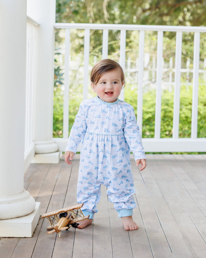 A toddler stands barefoot on a porch wearing a light blue long-sleeve jumpsuit with a small dinosaur print. A wooden toy airplane lies on the deck beside the child, with white railings and green foliage in the background, that store
