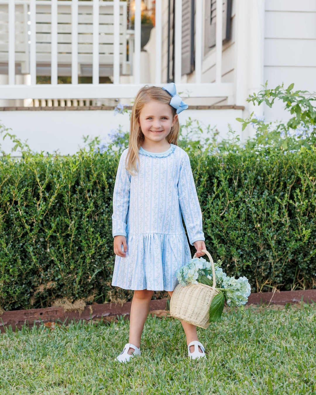 A young girl stands on a grassy lawn in front of a green hedge and a white house, wearing a light blue and white vertically striped long-sleeve dress and holding a woven basket of flowers. She has a light blue bow in her hair, that store
