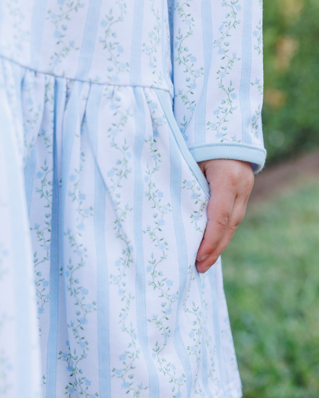 Close-up of a light blue and white vertically striped dress with a delicate green floral pattern, featuring a side pocket and a hand resting in the pocket, set against a blurred green outdoor background, that store