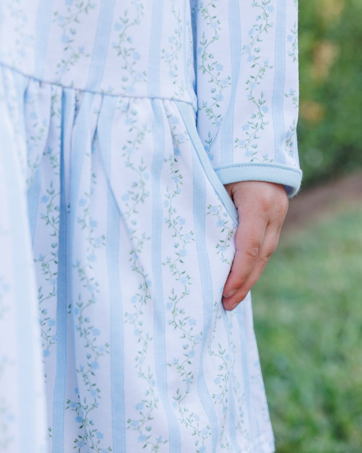 Close-up of a light blue and white vertically striped dress with a delicate green floral pattern, featuring a side pocket and a hand resting in the pocket, set against a blurred green outdoor background, that store