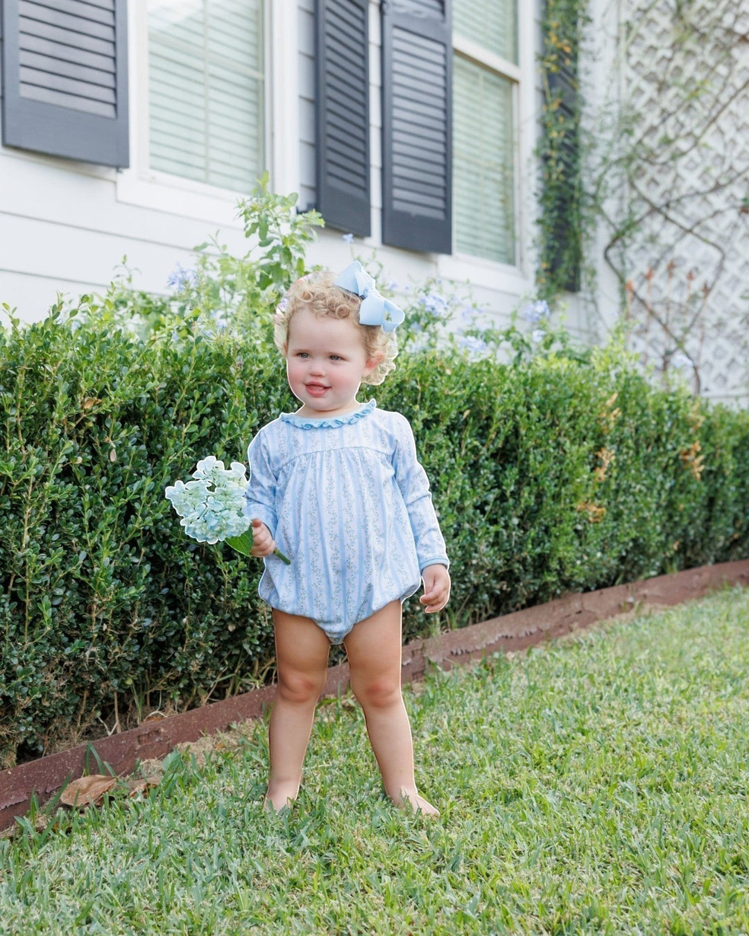 A toddler girl stands barefoot on a grassy lawn in front of a house, wearing a light blue and white vertically striped long-sleeve romper and holding a small bouquet of light blue hydrangeas. She has a matching light blue hair bow in her hair, that store
