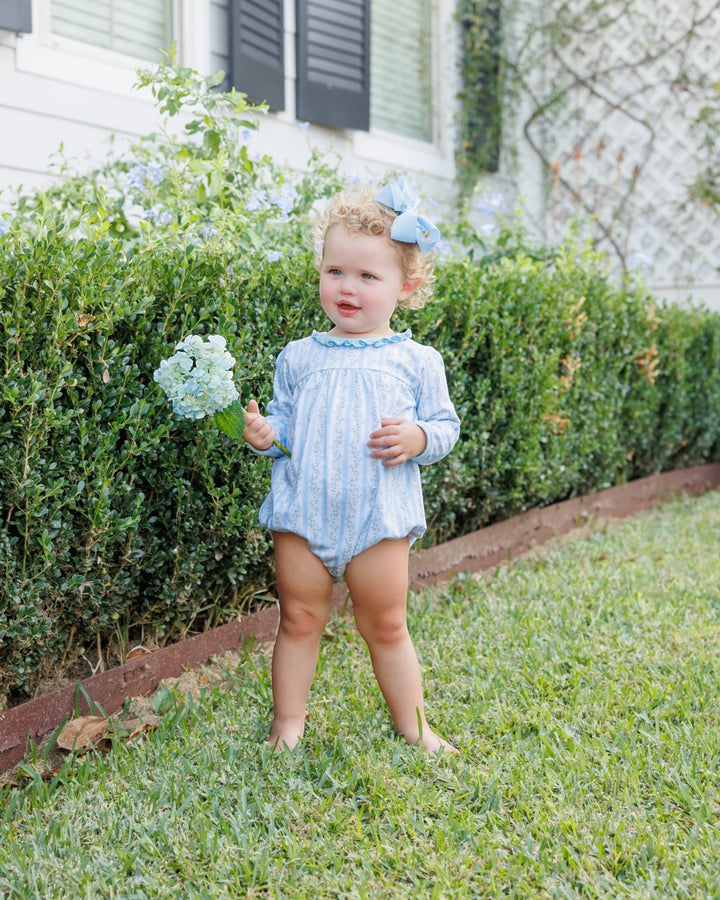 A toddler standing barefoot on a grassy lawn in front of a hedge, wearing a light blue and white vertically striped long-sleeve romper with a matching blue bow in their hair and holding a small bouquet of hydrangeas, that store