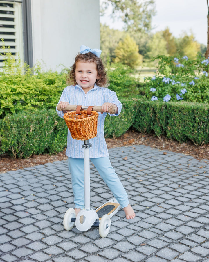 A young child stands barefoot on a paved outdoor path holding a white and wicker scooter with a front basket, with hedges and a house in the background, that store