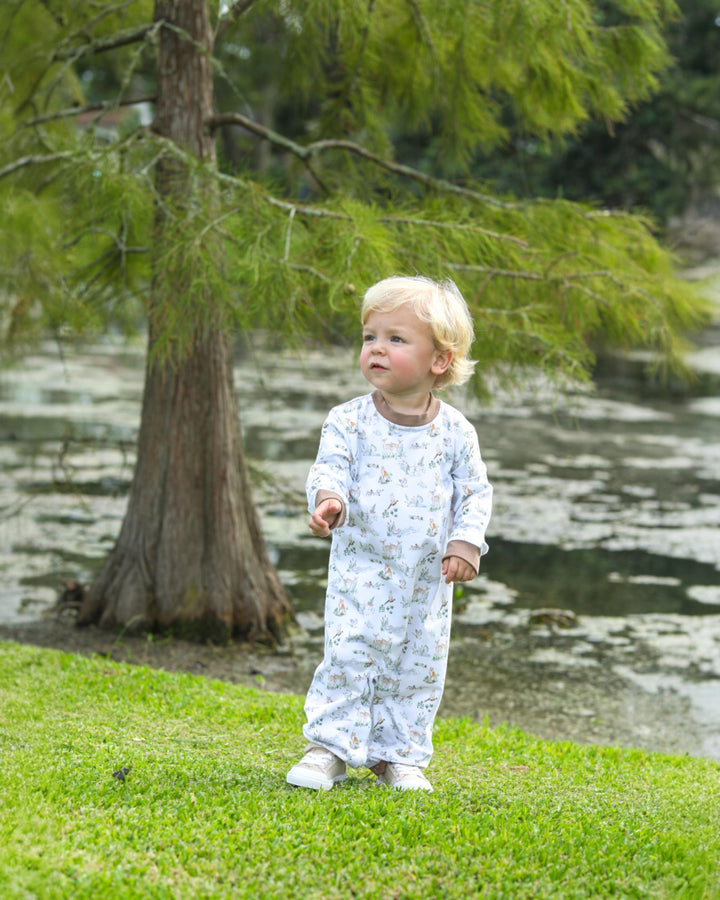 Child in a white onesie with blue patterns standing on grass near a tree and water.