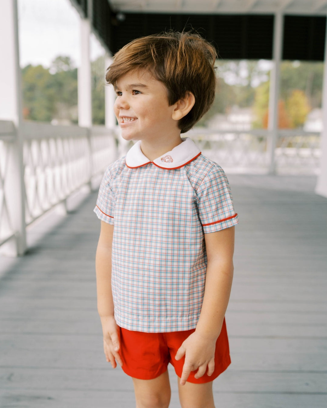 Child wearing a checkered shirt with a collar and red shorts on a wooden deck.