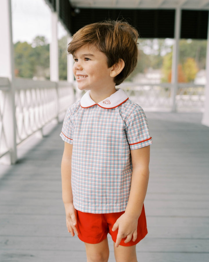 Child wearing a checkered shirt with a collar and red shorts on a wooden deck.