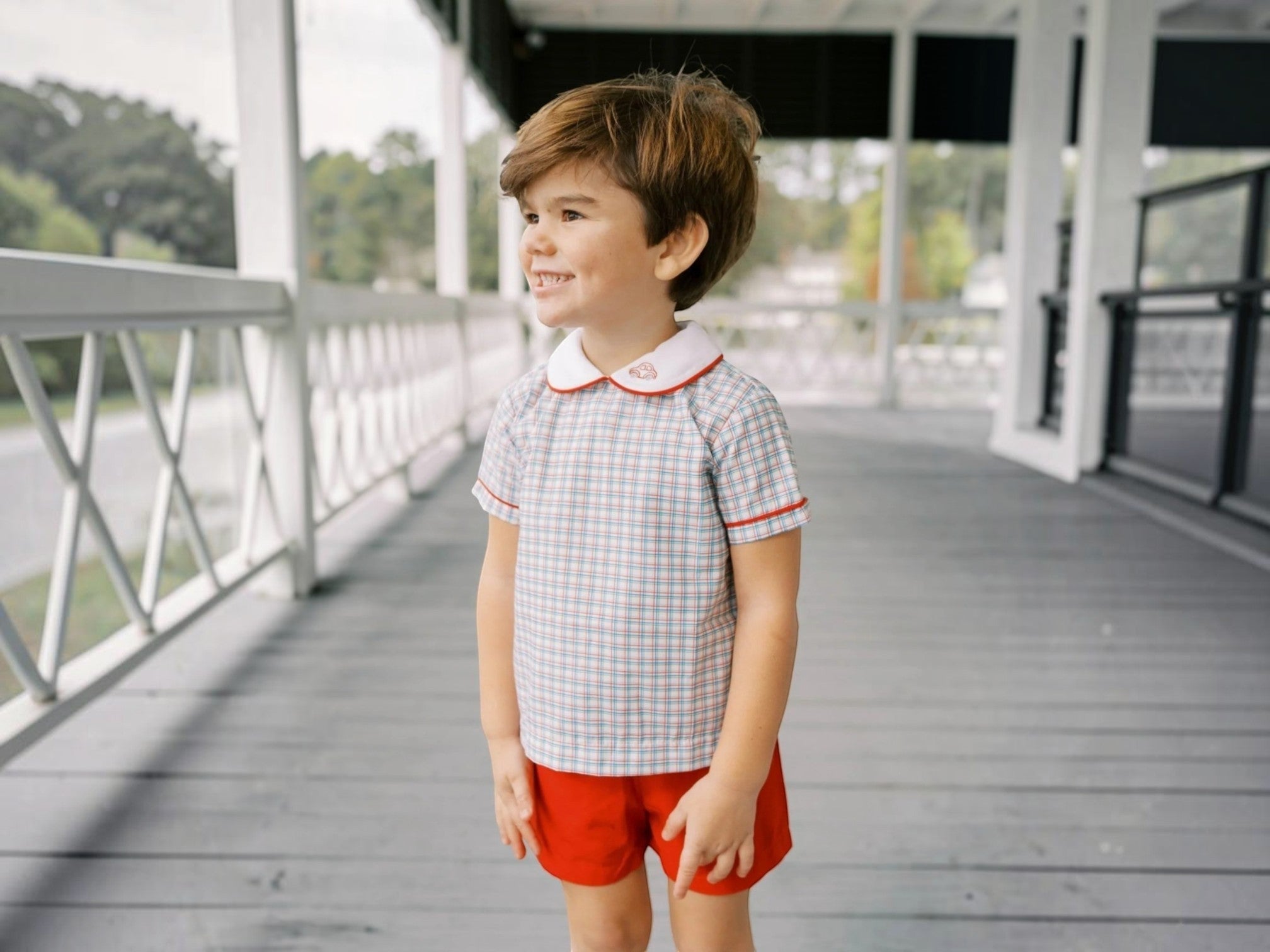 Young boy standing on a wooden deck wearing a checkered shirt and red shorts.