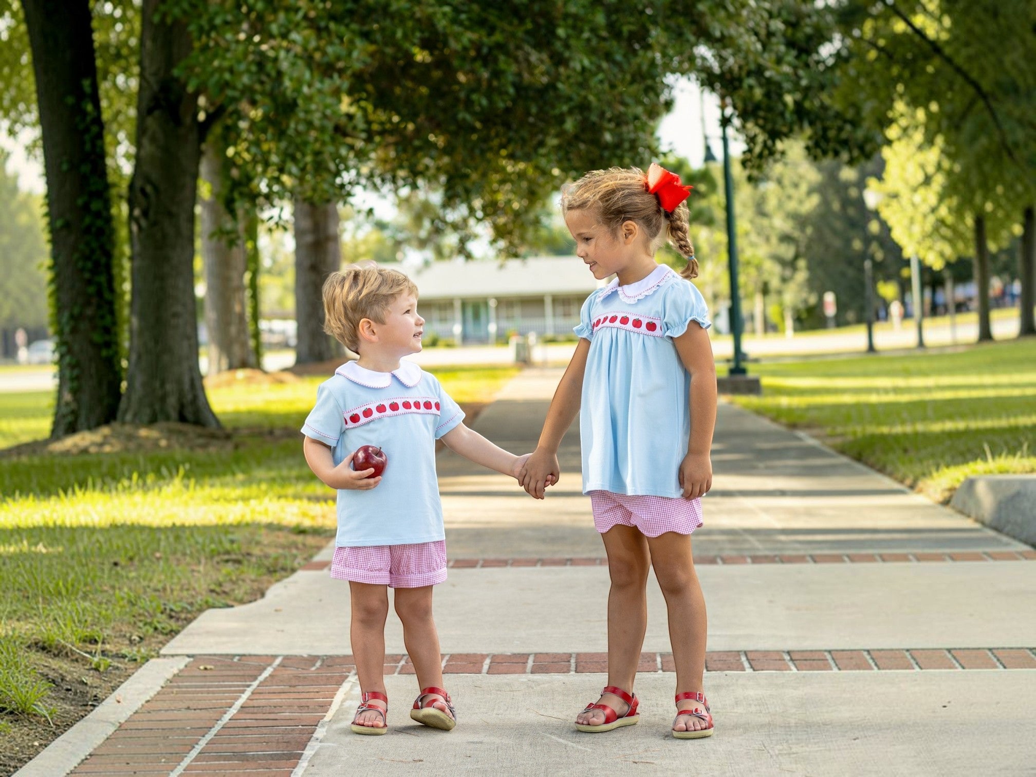 Two young girls standing on a paved path in a park, wearing matching light blue smocked dresses with red trim and pink shorts. One girl holds a red apple, that store
