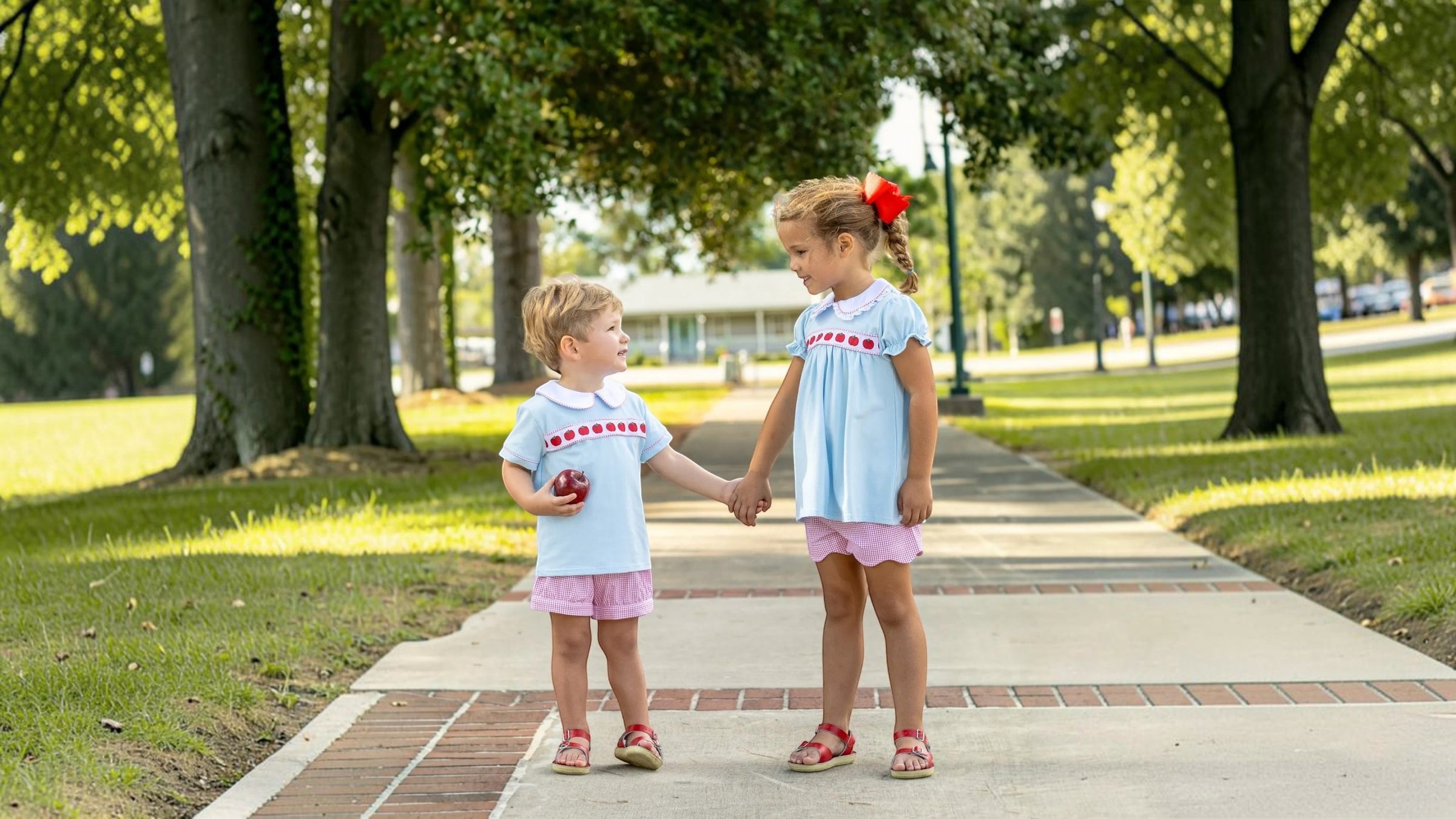 Two young girls standing on a paved path in a park, holding hands. Both are wearing matching light blue short-sleeve dresses with red text on the chest and pink shorts, that store