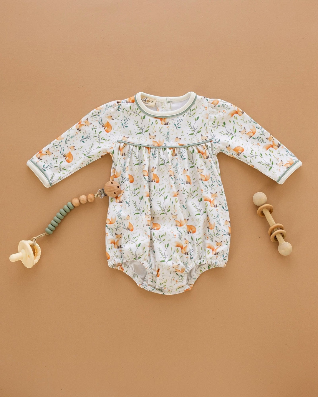 A baby long-sleeve bodysuit with an all-over fox and leaf print, styled in a flat lay on a warm tan background with a pacifier, wooden beads, and a wooden rattle, that store