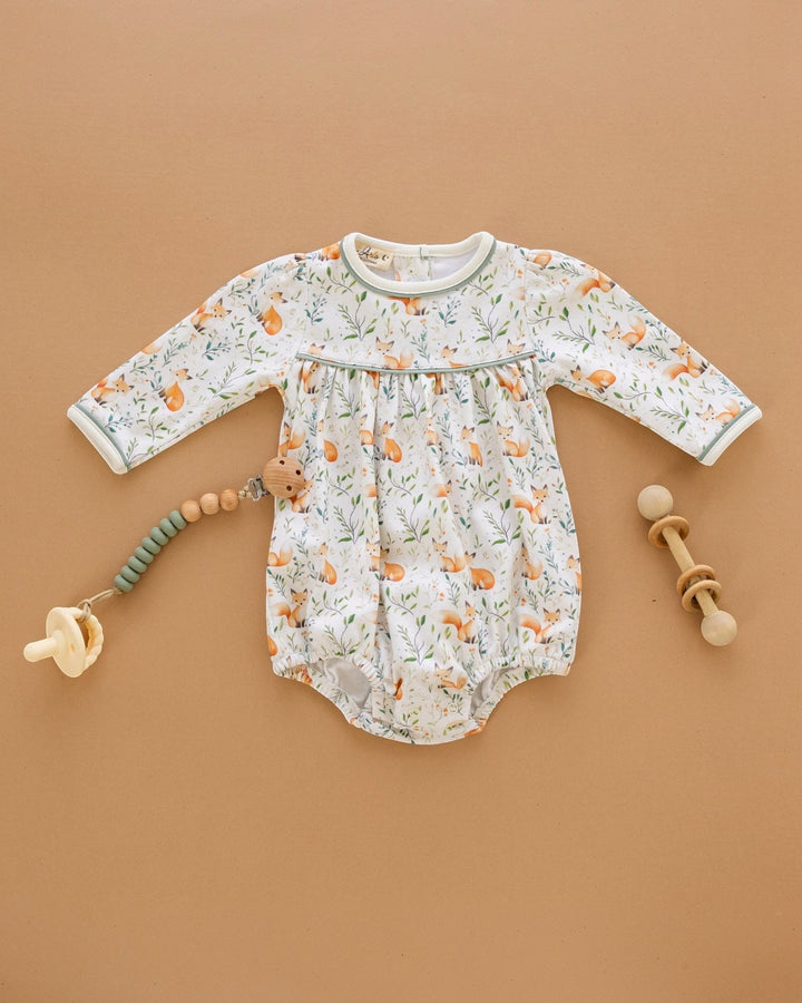 A baby long-sleeve bodysuit with an all-over fox and leaf print, styled in a flat lay on a warm tan background with a pacifier, wooden beads, and a wooden rattle, that store