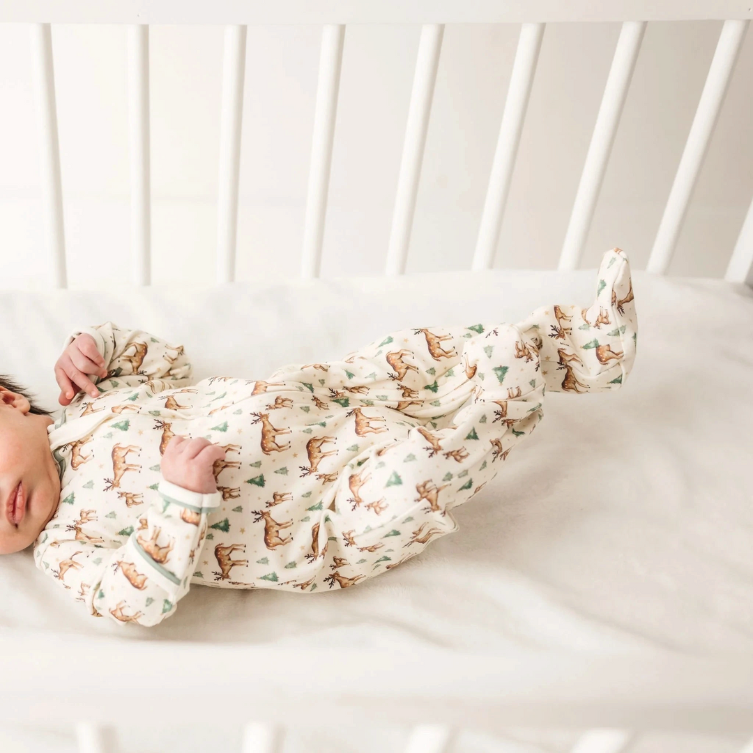 That Store baby footie in ivory Pima cotton with woodland deer print and moss green trim, worn by a baby in a crib.