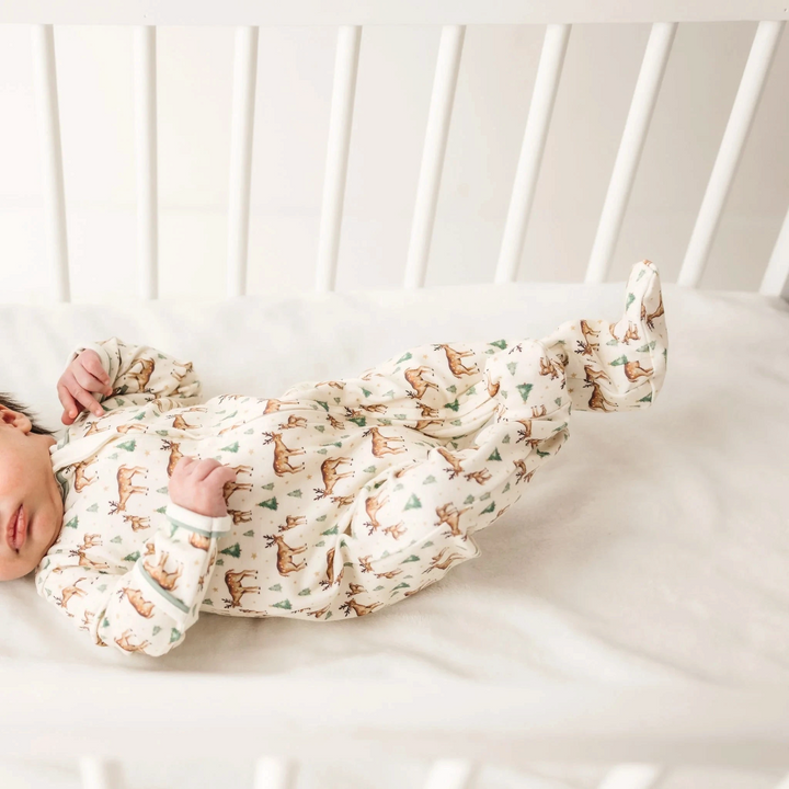 That Store baby footie in ivory Pima cotton with woodland deer print and moss green trim, worn by a baby in a crib.