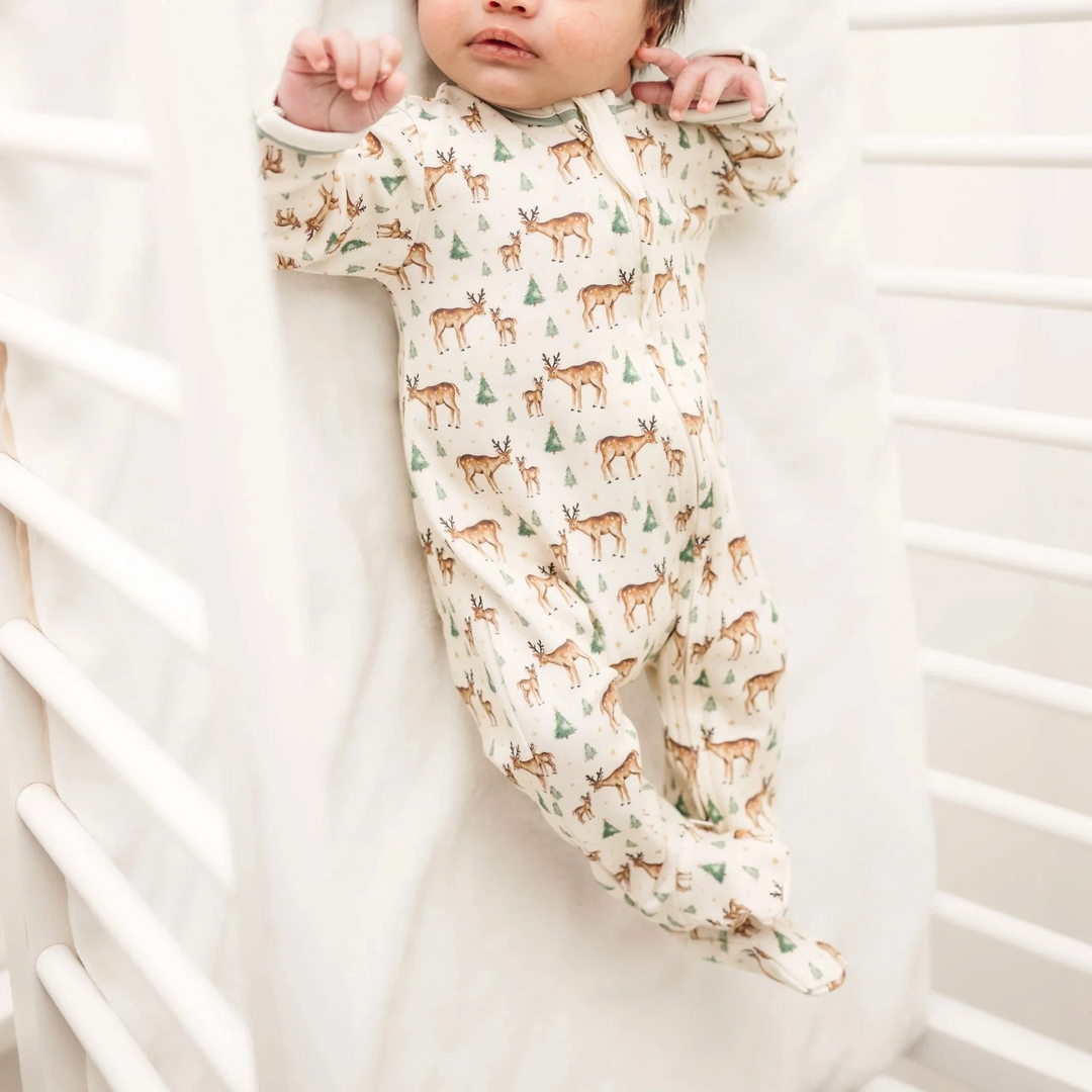 That Store baby footie in ivory Pima cotton with a woodland deer, fawn, and evergreen tree print and moss green trim.