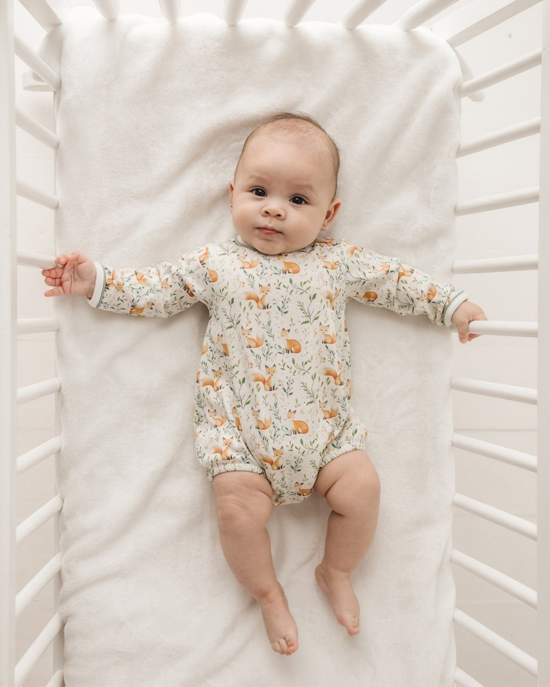 A baby lying on a white blanket inside a white crib, wearing a long-sleeve cream-colored romper with a delicate orange and green woodland animal print, that store