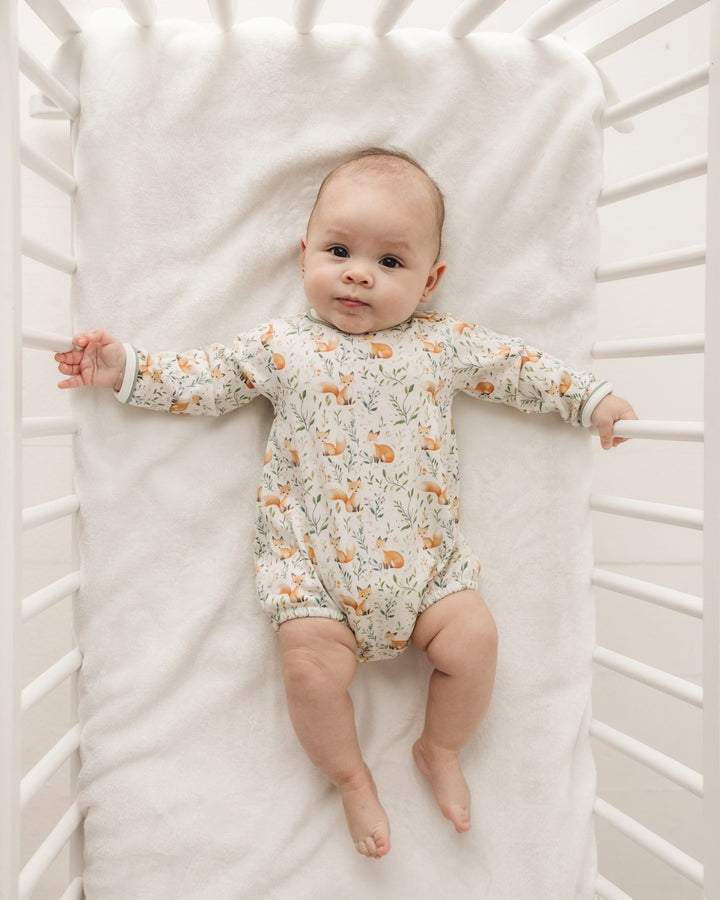 A baby lying on a white blanket inside a white crib, wearing a long-sleeve cream-colored romper with a delicate orange and green woodland animal print, that store