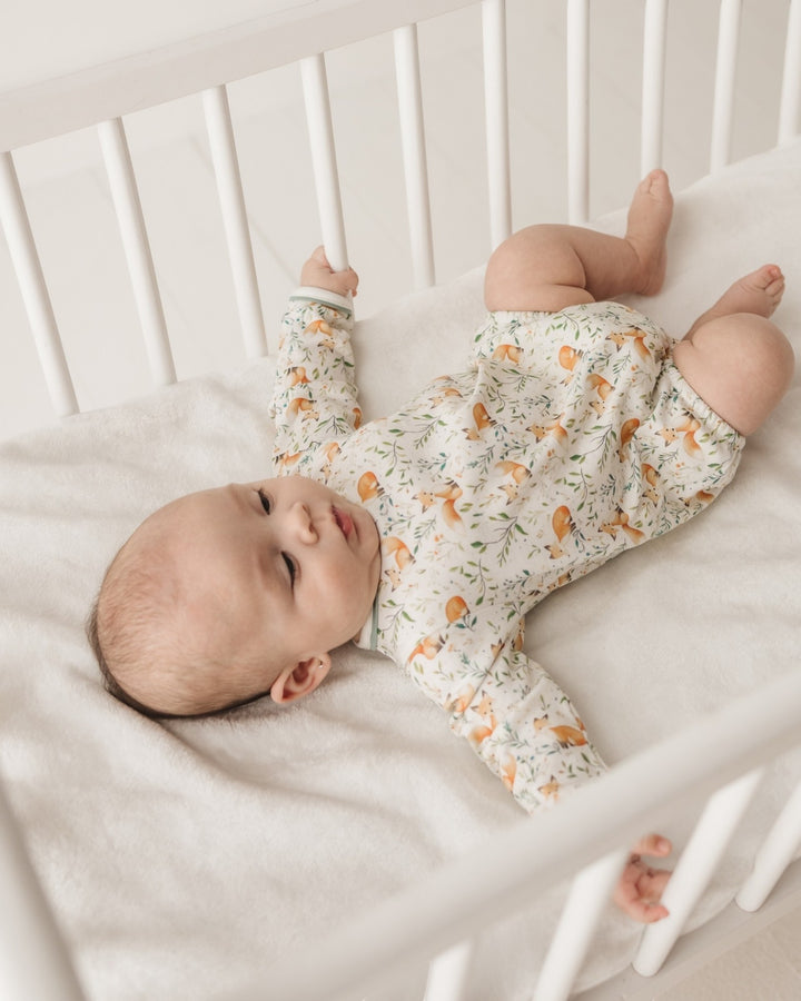 A baby lying on their back in a white wooden crib, wearing a long-sleeve cream-colored romper with a delicate green floral pattern and orange fox motifs, that store