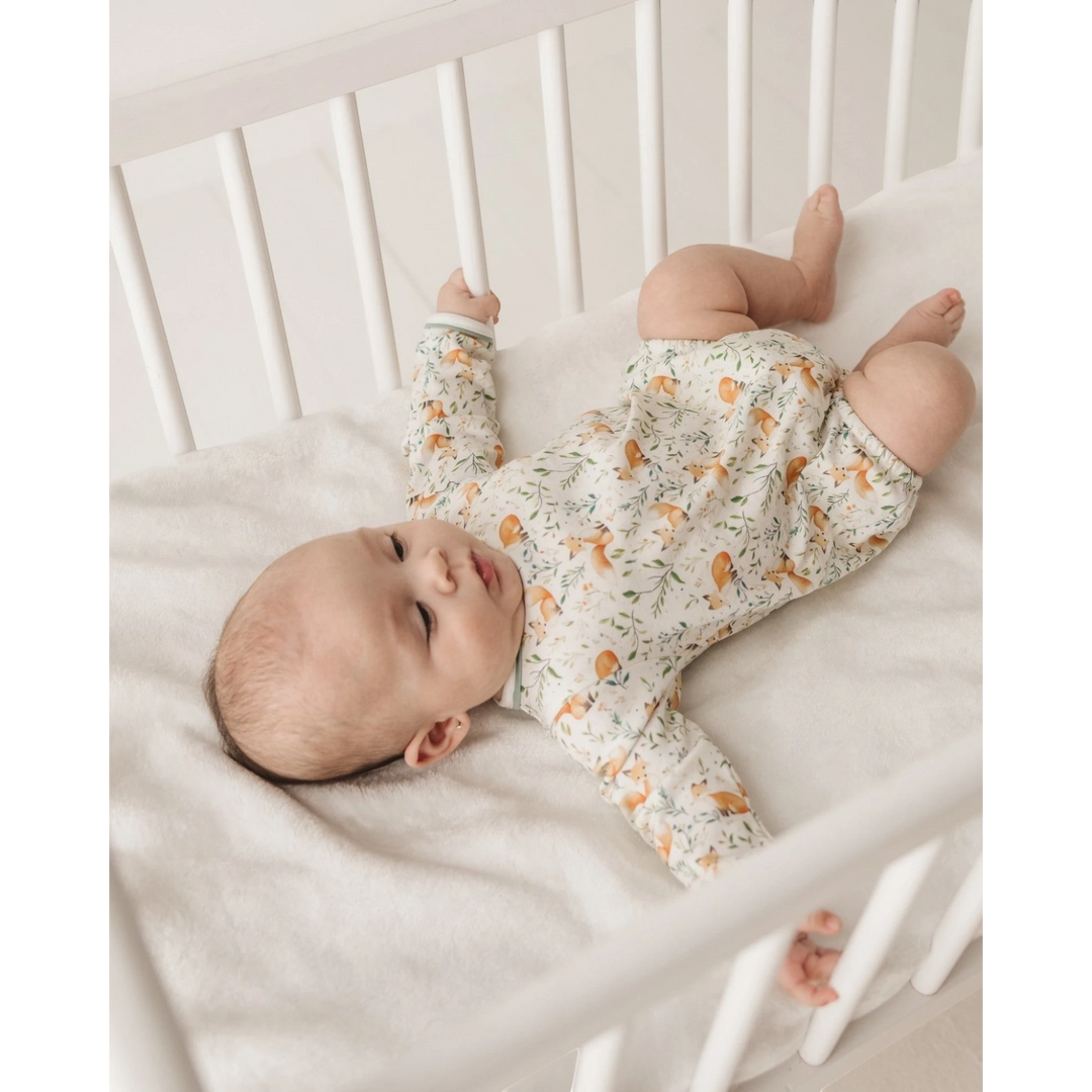 A baby lying on their back in a white wooden crib, wearing a long-sleeve cream-colored romper with a delicate green floral pattern and orange fox motifs, that store