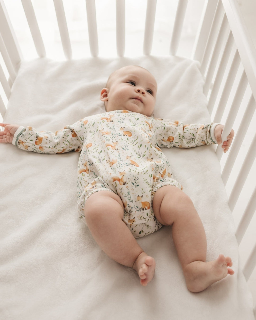 A baby lying on their back in a white wooden crib, wearing a long-sleeve cream-colored romper with a delicate orange and green animal print, that store