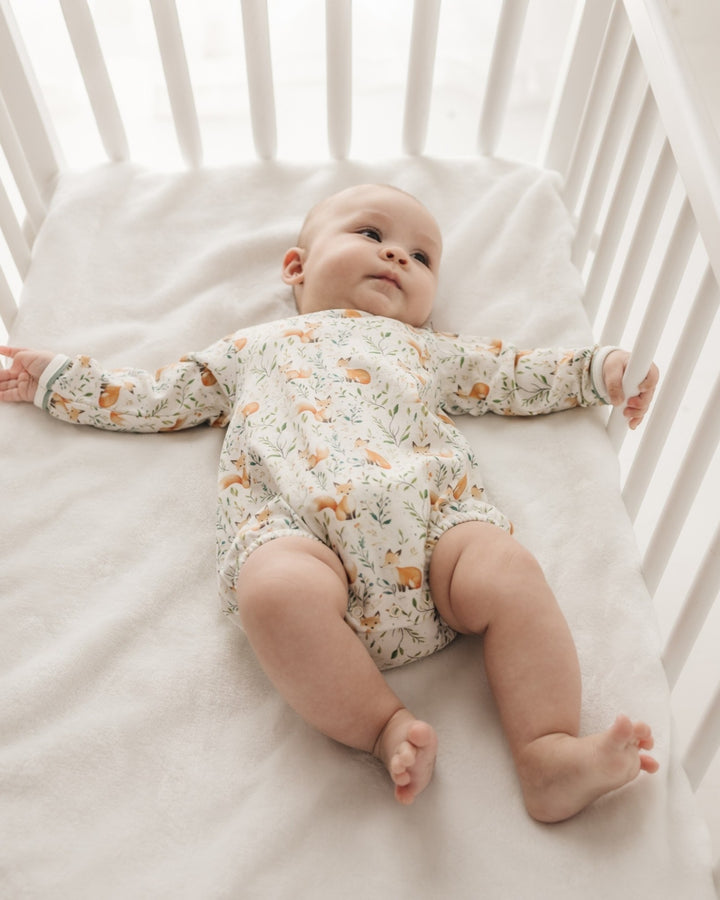 A baby lying on their back in a white wooden crib, wearing a long-sleeve cream-colored romper with a delicate orange and green animal print, that store